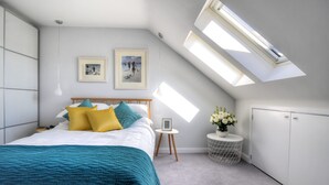 A bedroom with a sloped ceiling, skylight, bed with teal bedding, two framed pictures, bedside table with flowers, and a white wardrobe. - 3 Grove Mews (Seaton)