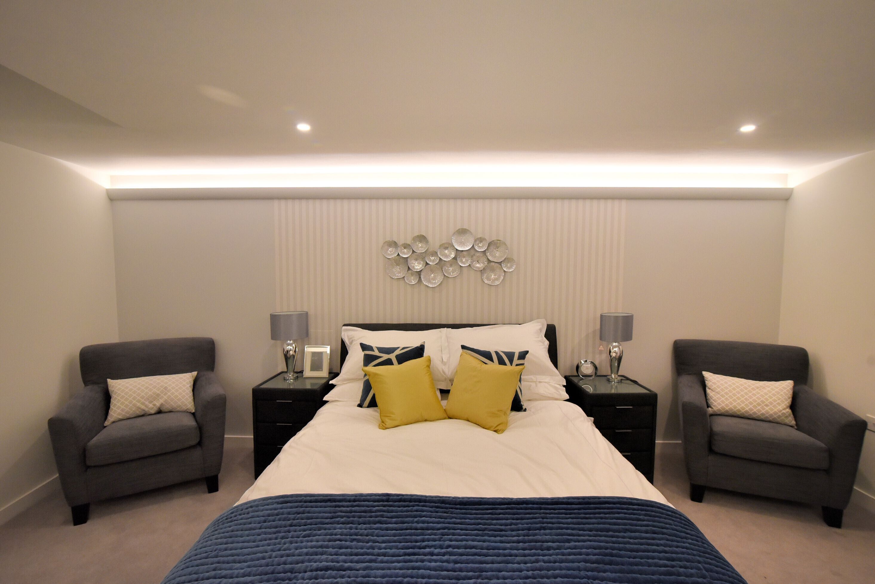 Quarto com uma cama grande, duas mesas de cabeceira, duas poltronas e um objeto decorativo na parede.