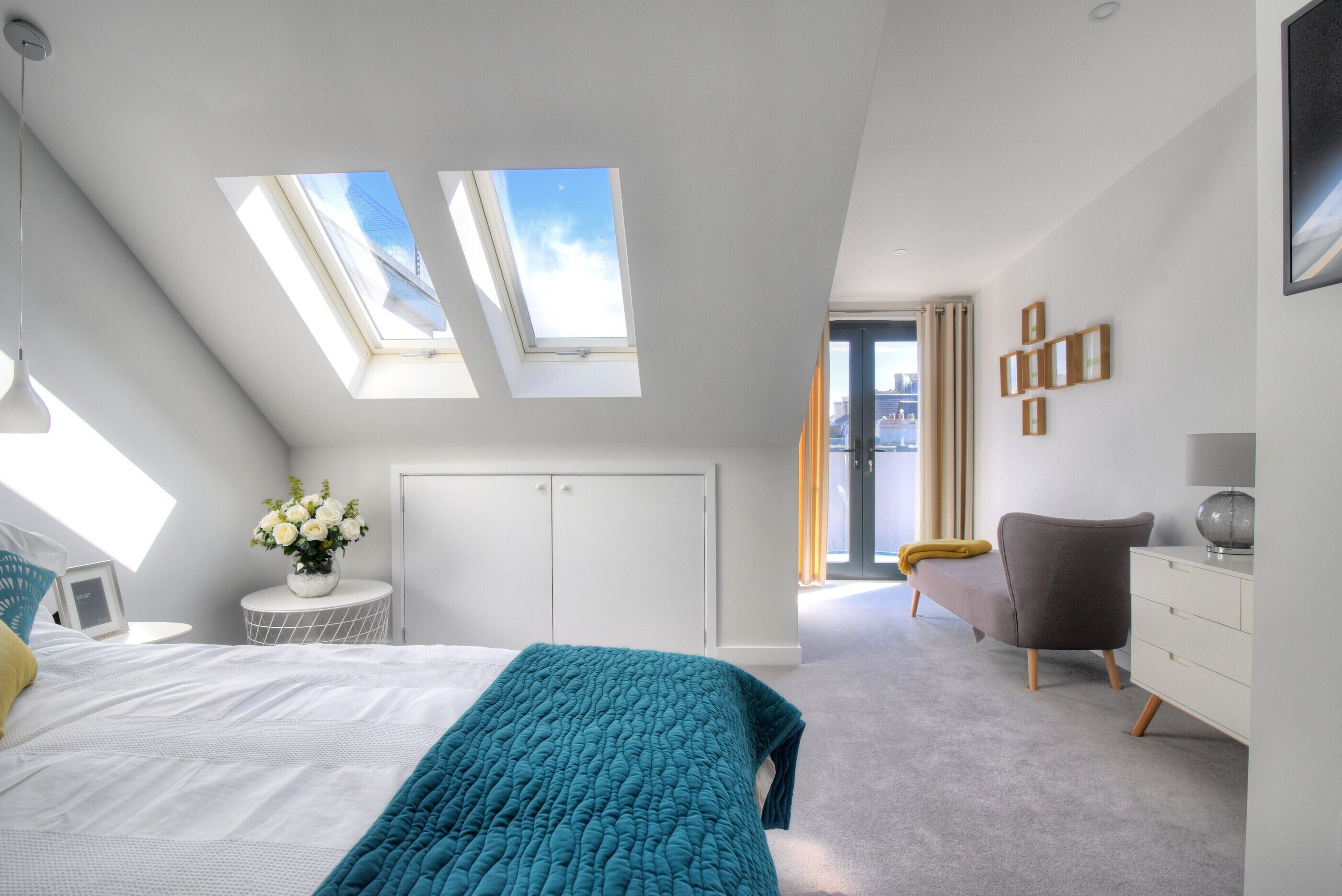 A modern bedroom with a skylight, a bed with a teal blanket, a bedside table with a vase of flowers, and a grey armchair.