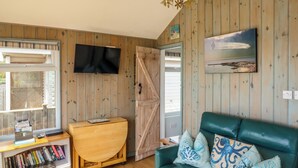 Una habitación acogedora con paredes de madera, un sofá de cuero verde, una mesita de madera con libros, un televisor montado en la pared y un ventanal grande.