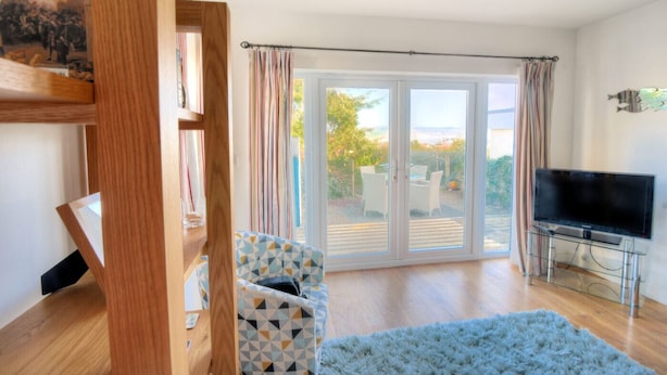 A living room with a TV, a sofa, a patterned armchair, and a large sliding glass door leading to an outdoor patio.
