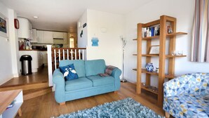 A living room with a blue sofa, a patterned rug, a wooden bookshelf, and a kitchen in the background.
