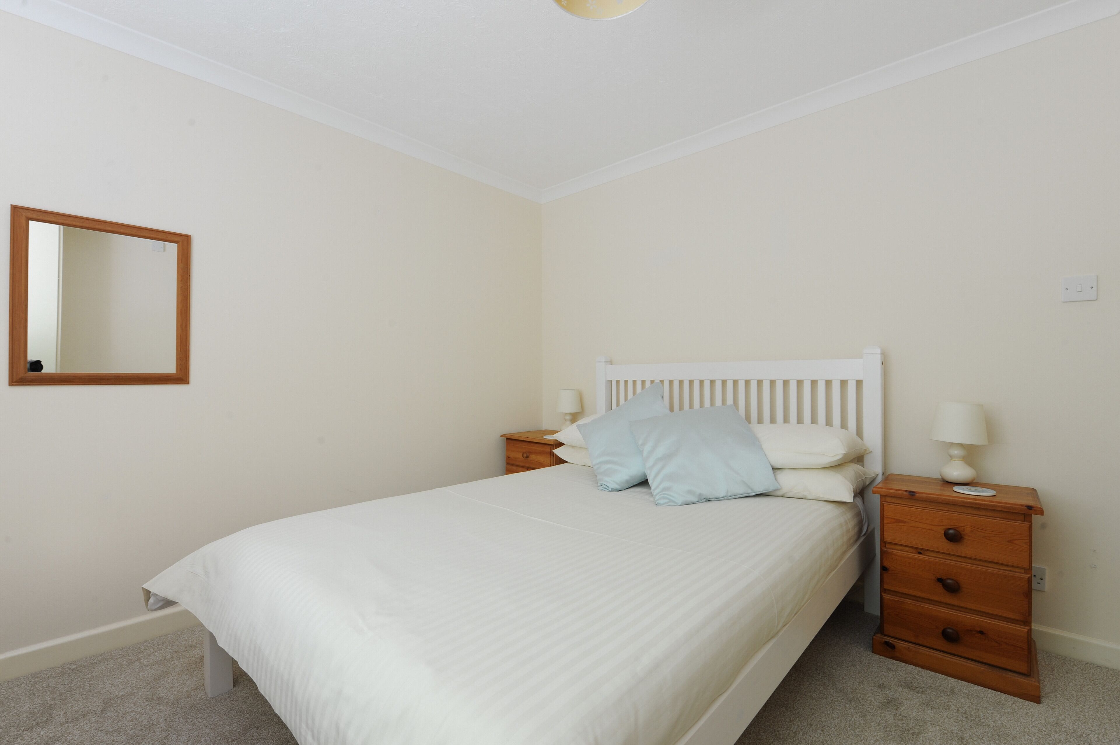 A bedroom with a bed, a nightstand, a drawer unit, a mirror, and a wall socket.