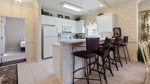 A modern kitchen with white cabinets, a central island with bar stools, and a dining area with a white table and chairs. - Stunning 3BR Condo Magnolia Point (Myrtle Beach)