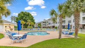 A pool area with lounge chairs and umbrellas, surrounded by palm trees and a building with balconies. - Stunning 3BR Condo Magnolia Point (Myrtle Beach)