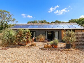 Un bâtiment moderne doté d’un toit recouvert de panneaux solaires, entouré d’une allée en gravier et agrémenté de différents pots de plantes.