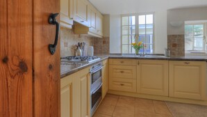 A kitchen with beige cabinets, a stove, and a window with flowers. - Tudor Cottage (Lyme Regis)
