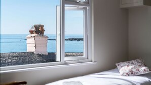 Une chambre avec vue sur la mer, visible par une fenêtre, un lit agrémenté d’un coussin à motifs floraux, et une structure en pierre à l’extérieur.