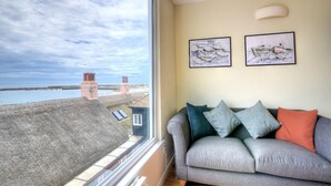 Un salon avec un canapé gris, deux coussins et une fenêtre donnant sur une zone côtière, avec des bâtiments et une vue sur la mer.