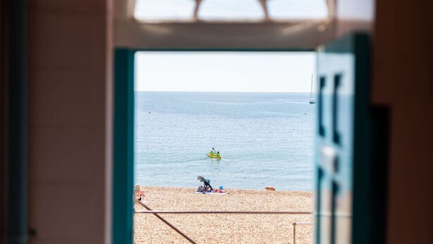 On aperçoit, à travers une porte ouverte, une plage où une personne fait du kayak et une autre est assise sur le sable.