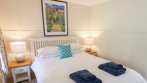 Una cama bien hecha con ropa de cama blanca, dos toallas azules y una almohada estampada en color aguamarina. Hay dos lámparas de noche sobre mesitas de madera y un cuadro enmarcado sobre la cama.