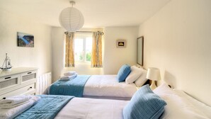A bedroom with two beds, a dresser, a window with curtains, a hanging light fixture, and a small framed picture on the wall. - Smugglers Cottage (Seaton)