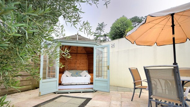 A cozy outdoor seating area with a wooden deck, a small open-sided structure with a bed, and a table with chairs. - Smugglers Cottage (Seaton)