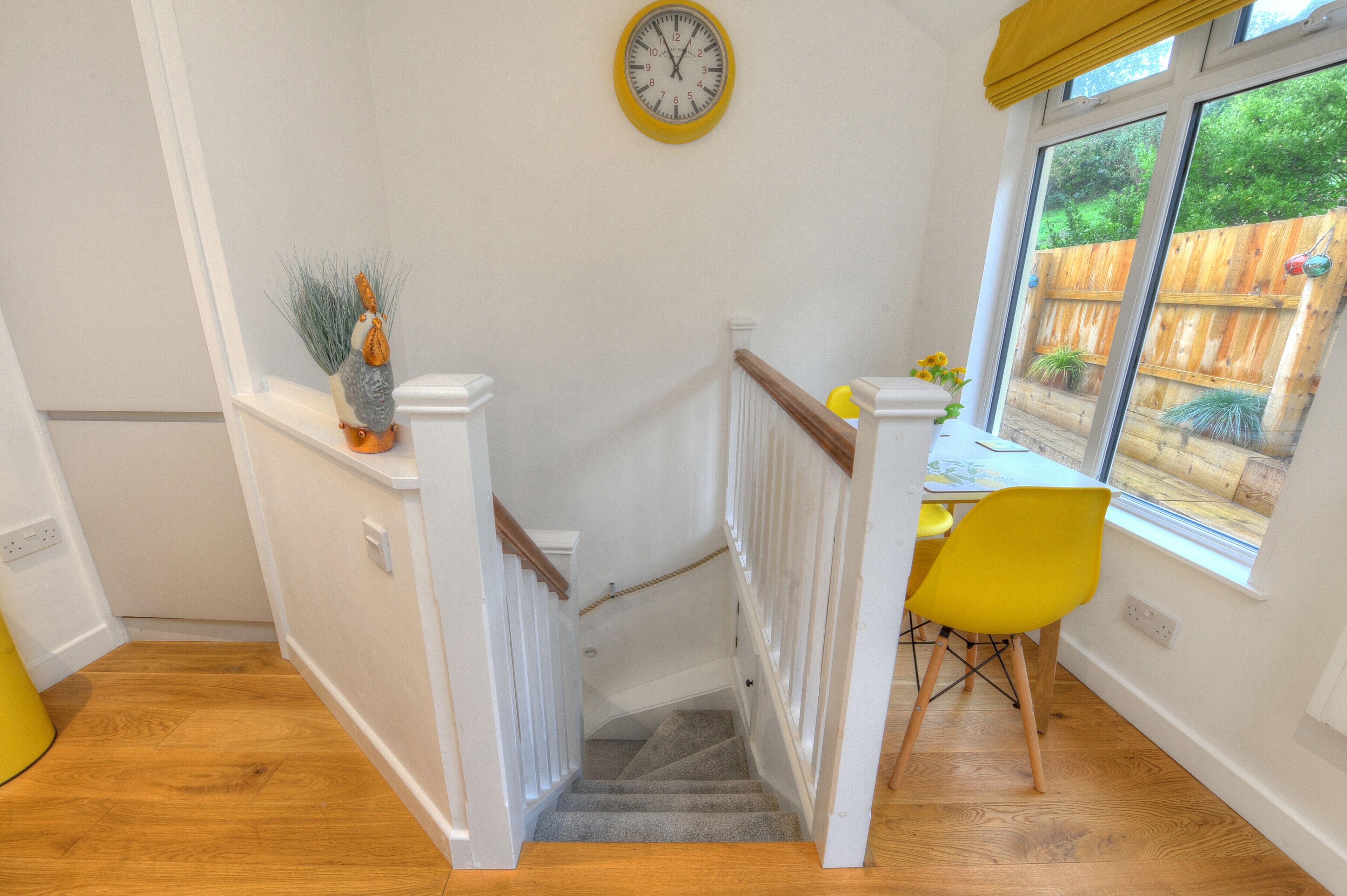 A staircase with a wooden handrail, a yellow wall clock, and a view of a wooden fence and greenery outside.