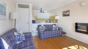 A living room with two blue plaid sofas, a white door, a kitchen area with a microwave, and a TV mounted on the wall. - 6 Bay View Court (Lyme Regis)