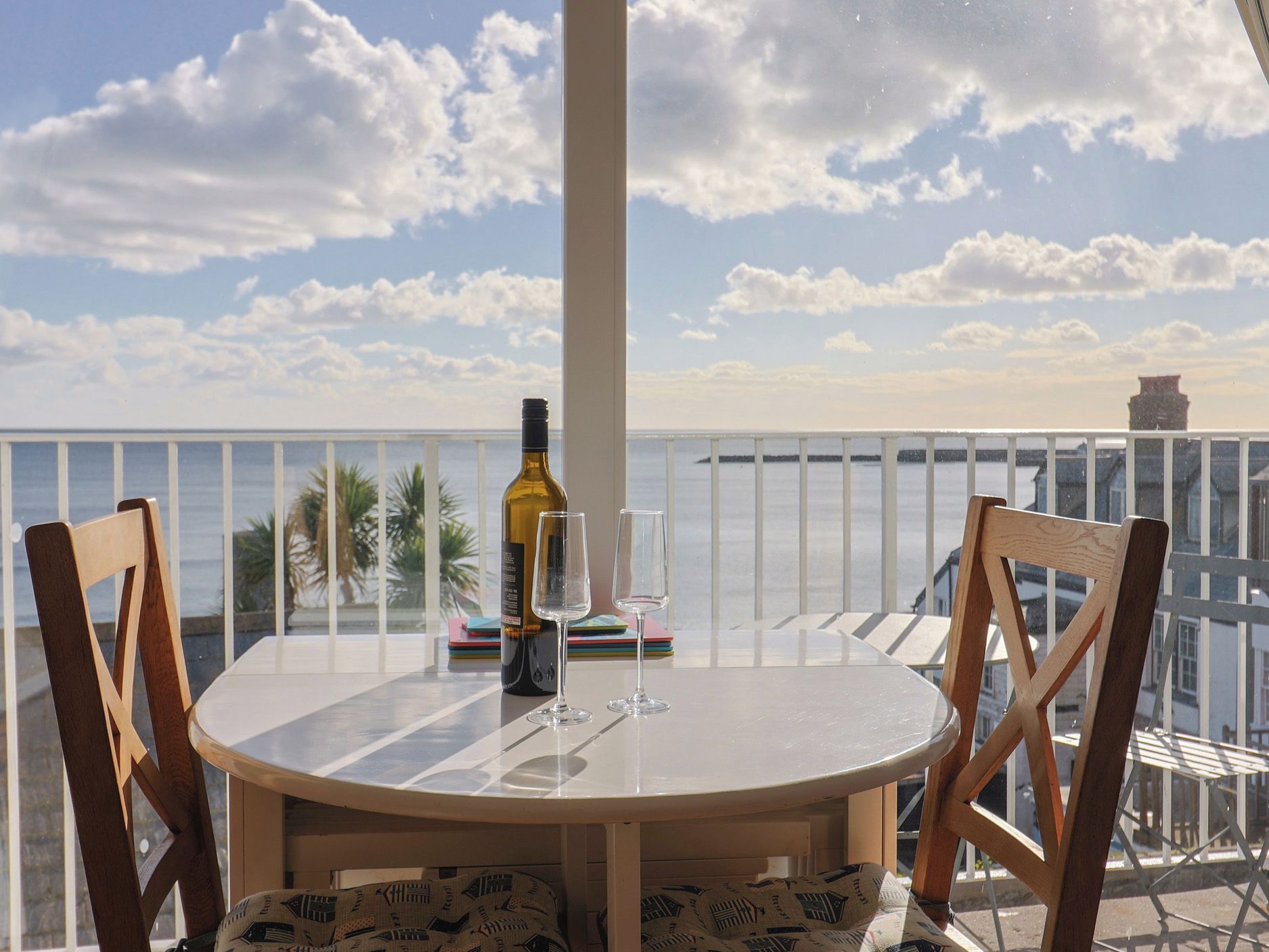 A round table with two chairs, a bottle of wine, and two glasses on a balcony overlooking the sea.