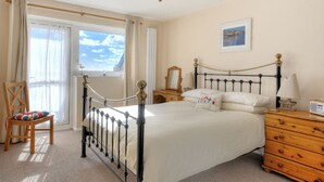 A bedroom with a bed, a wooden chair, a dresser, a nightstand, a lamp, and a window with curtains. - 6 Bay View Court (Lyme Regis)