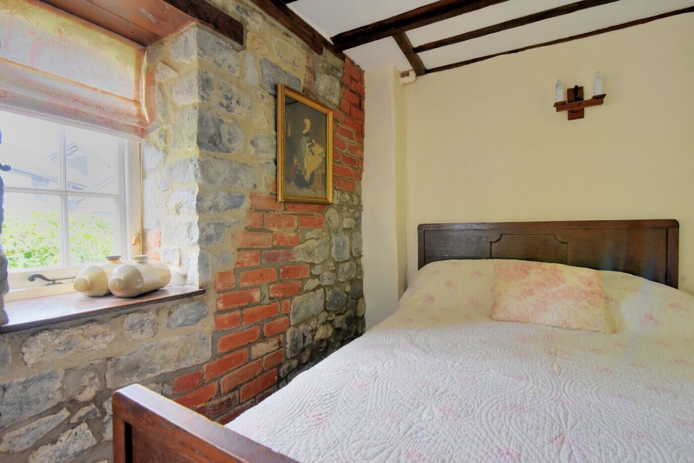 A bedroom with a stone wall, a wooden headboard, a window with bottles on the sill, and a painting on the wall.