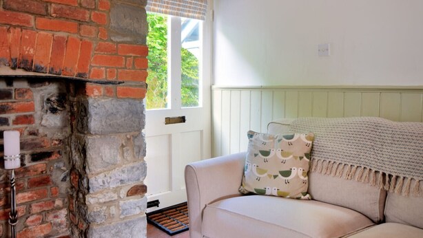 A cozy room with a brick fireplace, a white door, and a beige sofa with patterned cushions. - Lavender Cottage (Lyme Regis)