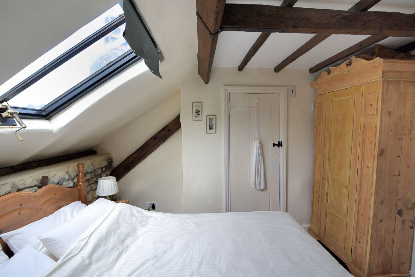 A bedroom with a skylight, a wooden wardrobe, a bed with white linens, and a white door.