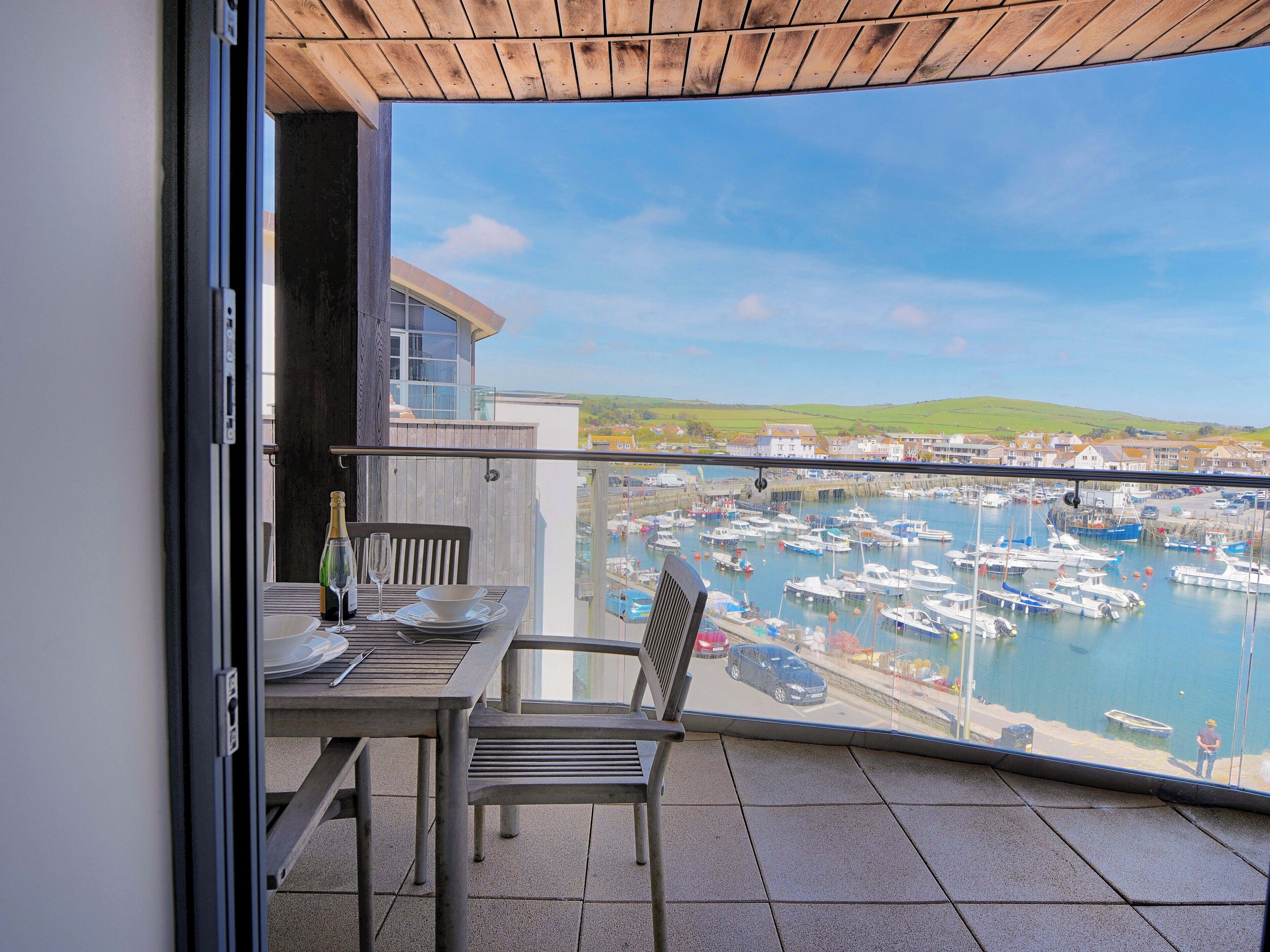 Un balcon avec une table et des chaises, donnant sur une marina où l’on aperçoit des bateaux et des maisons au loin.