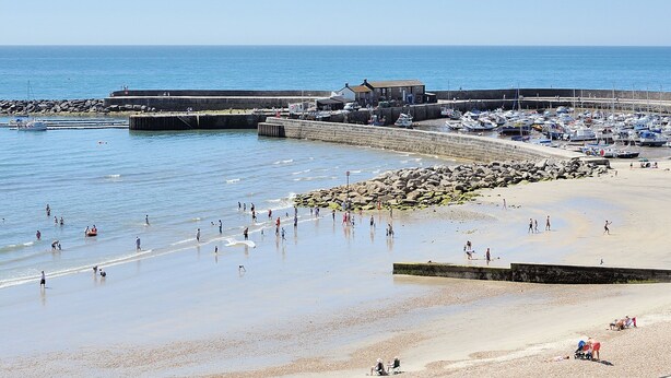 Une plage côtière avec une digue rocheuse, un port de plaisance avec des bateaux et un bâtiment doté d’un toit en pente.