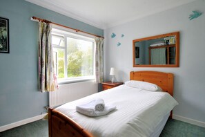 A bedroom with a bed, a window with curtains, a bedside table with a lamp, and a mirror on the wall. - Rooks Acre (Lyme Regis)