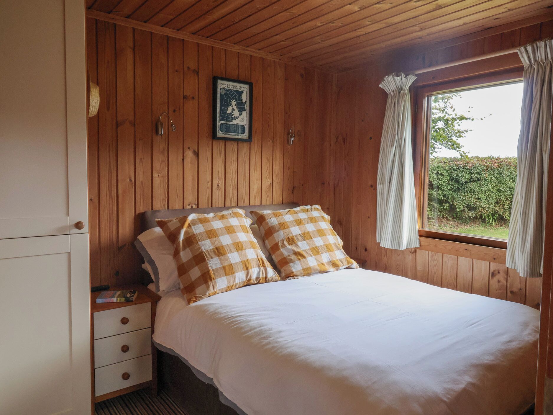 Habitación con paneles de madera, una cama, una mesita de noche, un aparador y una ventana con cortinas.