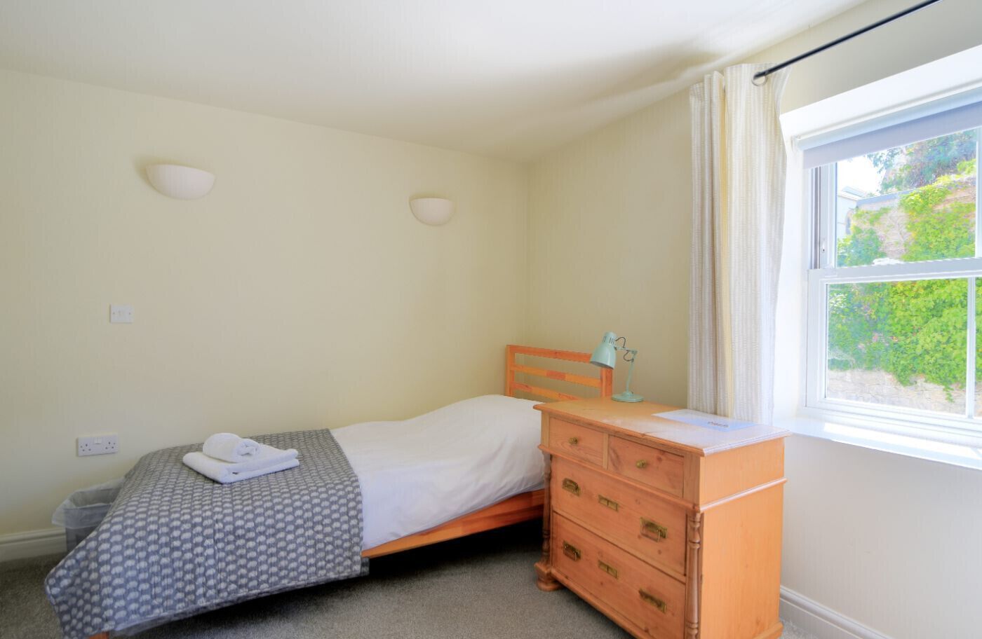 A small, neatly arranged bedroom with a single bed, a wooden dresser with drawers, a bedside lamp, and a window with a view of greenery.