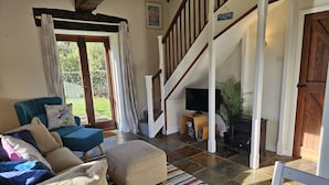 A cozy living room with a sofa, ottoman, and a glass of wine on a wooden table. A TV is mounted on a wooden stand, and a staircase leads to another room. - The Cider House (Axminster)