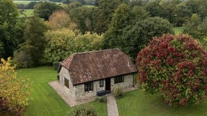 A stone house with a sloped roof, surrounded by a well-maintained lawn and dense trees. - The Cider House (Axminster)