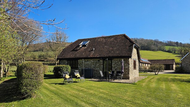 A stone building with a sloped roof, a covered patio with outdoor seating, and a view of a green hillside. - The Cider House (Axminster)