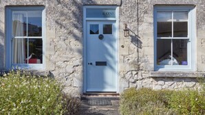 A stone building with a white door labeled "LONE," two windows with white frames, and a small garden area with bushes. - Lavender Cottage (Seaton)