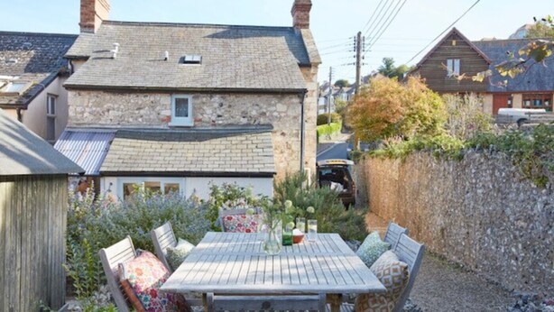 A stone house with a tiled roof, a wooden table with chairs, and a garden with various plants. - Lavender Cottage (Seaton)