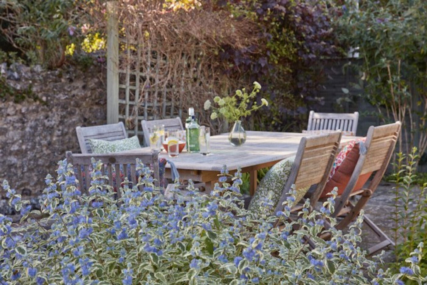 Una mesa de madera para comedor al aire libre con sillas, rodeada por un jardín de flores azules.