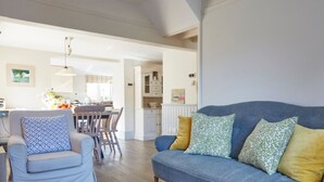 A living room with a sofa and armchair, a dining area with chairs, a kitchen in the background, and a large window. - Lavender Cottage (Seaton)
