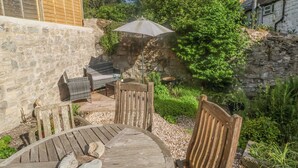 A patio with a round wooden table and chairs, a stone wall, and a small garden area with a bird bath and greenery. - Coombe Street Cottage (Lyme Regis)