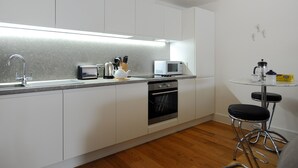 A modern kitchen with white cabinets, a stainless steel sink, a microwave, a kettle, and a coffee maker. There is a small round table with two stools. - Seashell (Lyme Regis)