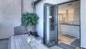 An outdoor patio with a wooden table, two potted plants, and a sliding glass door leading to a kitchen area. - No. 2, 26 Broad Street (Lyme Regis)