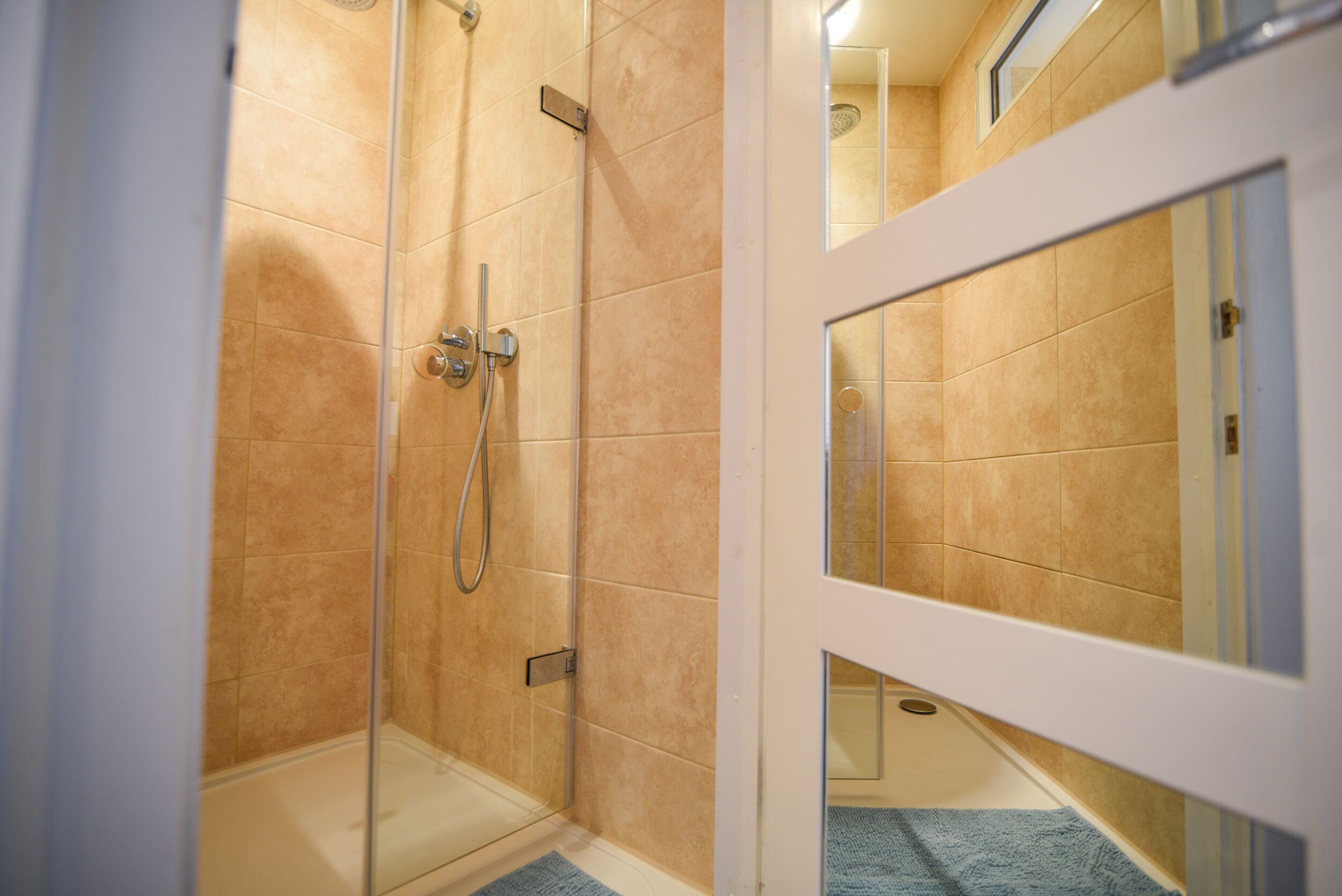A glass-enclosed shower with beige tiles, a white door frame, and a blue towel on the floor.