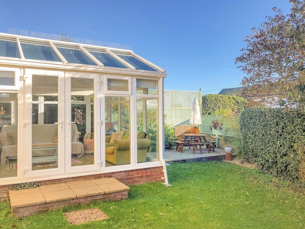 Un invernadero con techo de cristal, base de ladrillo y zona de patio con mobiliario exterior.