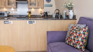 A modern kitchen with wooden cabinets, a granite countertop, and stainless steel appliances. There is a purple sofa with a colorful geometric pillow and a vase with flowers on the counter. - 2 Driftwood (Bridport)