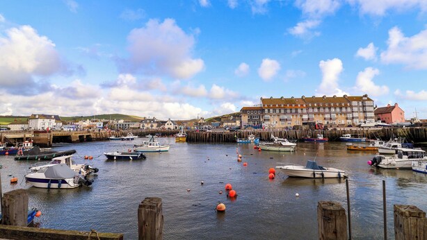 A harbor with boats docked, wooden piers, and a row of buildings in the background. - 2 Driftwood (Bridport)