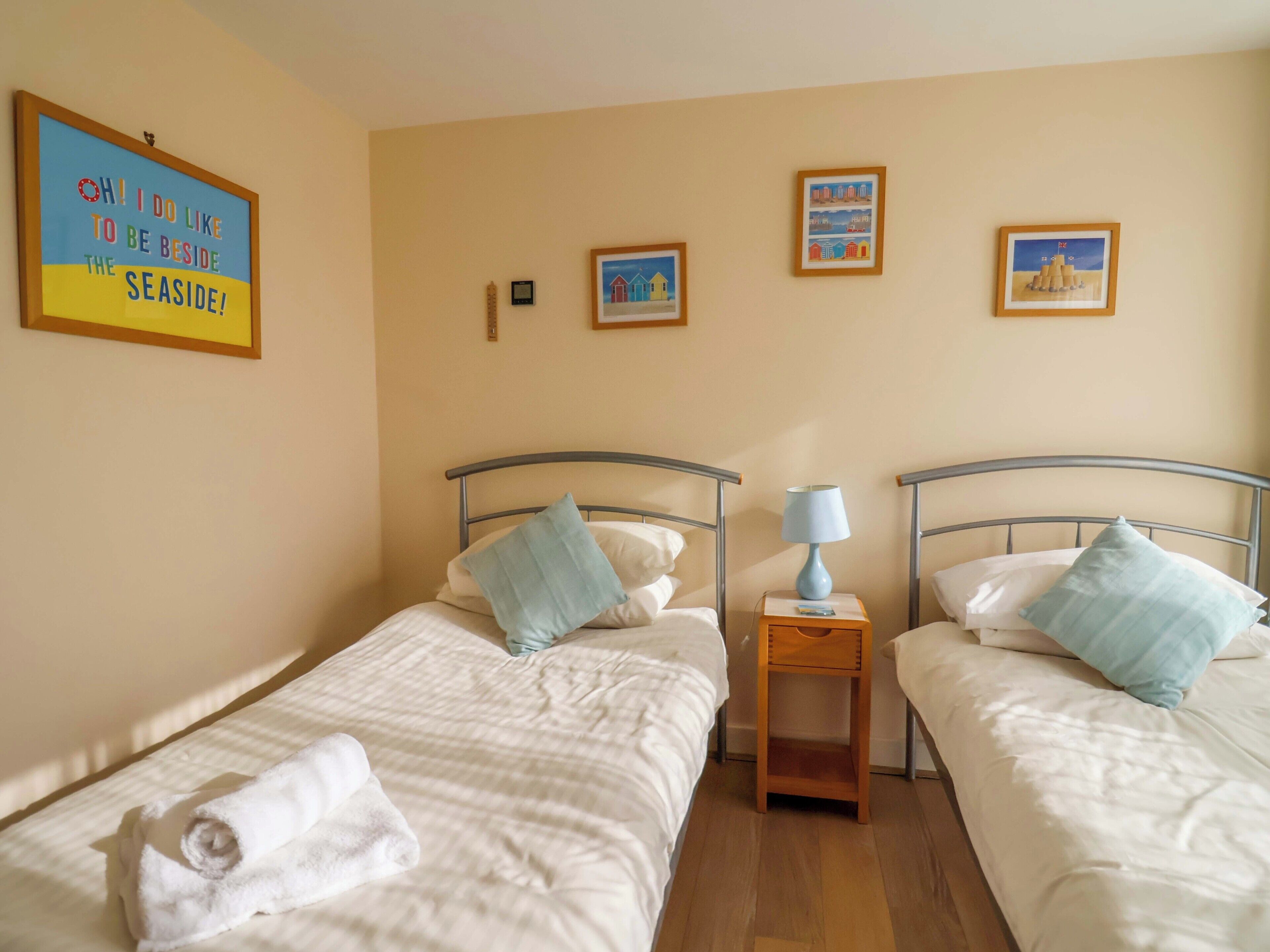 A room with two beds, each with a pillow and a rolled towel. There are framed pictures on the wall and a small table with a lamp between the beds.
