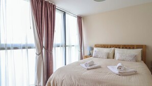A hotel room with a large bed, a nightstand with a lamp, and a view through the window. - 2 Driftwood (Bridport)