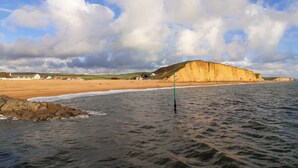 A beach with a prominent cliff, calm sea, and a partly cloudy sky. - 2 Driftwood (Bridport)