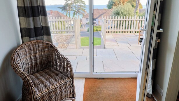 Una silla de mimbre en una habitación con un ventanal corredero que da a un patio con una cerca blanca y vistas a casas y árboles.
