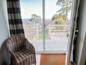 A wicker chair in a room with a sliding glass door leading to a patio with a white fence and a view of houses and trees. - Westhaven (Lyme Regis)