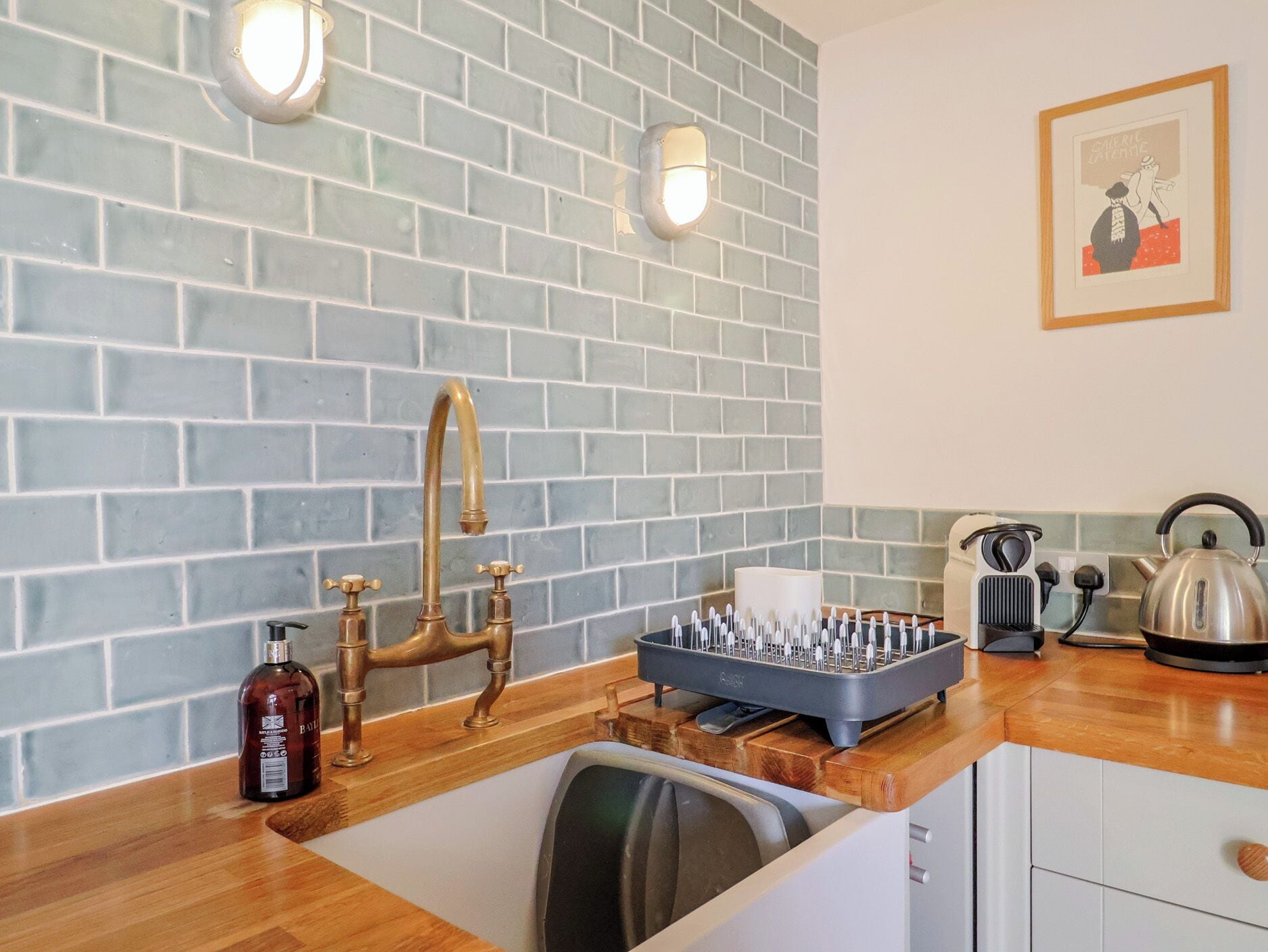 A kitchen with a blue-tiled backsplash, a brass faucet, a sink, a dish rack, a kettle, and a bottle of hand soap.