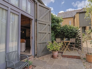 A patio area with a table and chairs, a wooden fence, and a large open garage door. - The Stone Barn (Bridport)